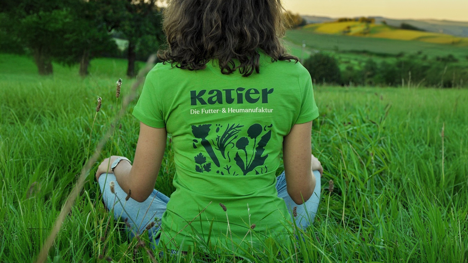 Frau sitzt im Gras, auf der Rückseite ihres T-Shirts ist das Katier Logo und Pattern zu sehen.